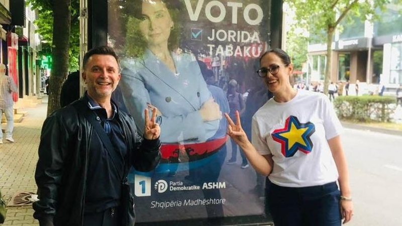 Erion Braçe dhe Delina Ibrahimaj pozojnë me dy gishtat lartë para posterit elektoral të Jorida Tabakut